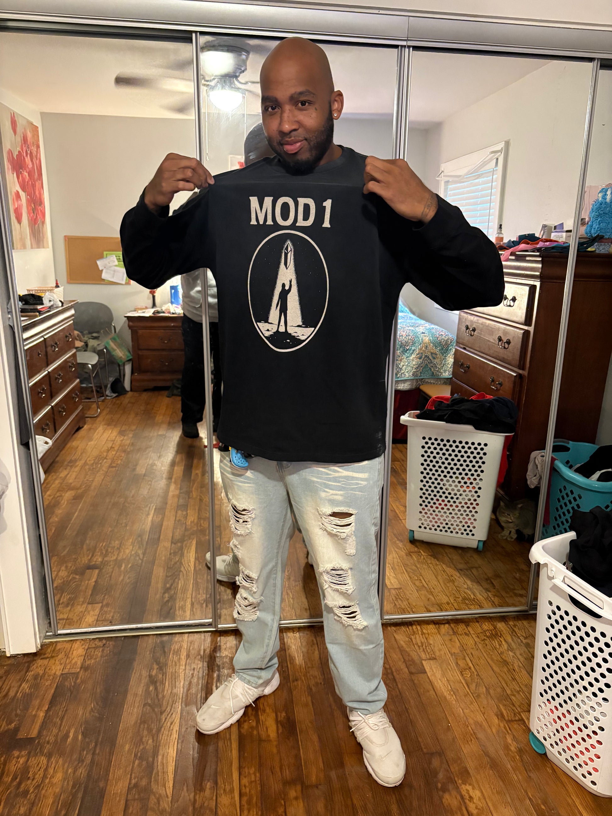 Man wearing a black acid washed MOD 1 Prototype Awakening Emblem sweatshirt with 'MOD 1' logo in a room with wooden floor and sliding glass door.