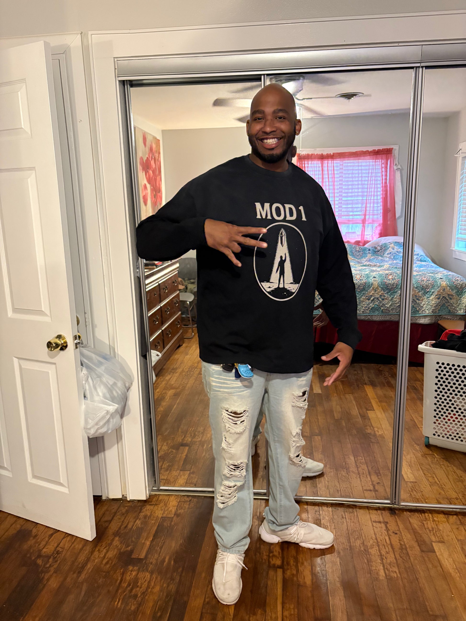 Man wearing a black acid washed MOD 1 Prototype Awakening Emblem sweatshirt with 'MOD 1' logo in a room with wooden floor and sliding glass door.